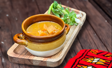 "A bowl of butternut squash soup with radish slices and pumpkin seeds, topped with cream. Served in a white bowl on a wooden surface, with a glass of red wine on the side.
