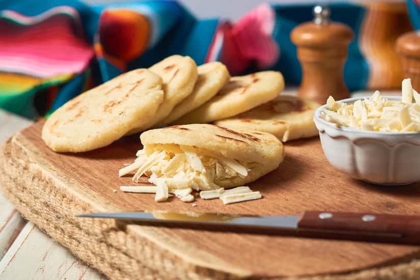 Arepas with shredded cheese on a wooden cutting board.
