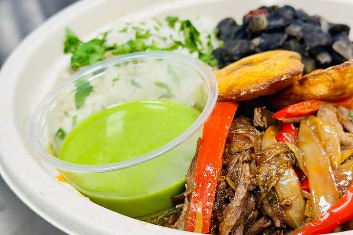Colorful Latin American meal with rice, black beans, plantains, beef, and green sauce.