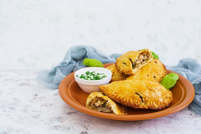 Baked empanadas on a terracotta plate with a bowl of dipping sauce, garnished with chopped herbs and green leaves.